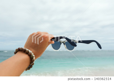A person holds sunglasses while standing by the ocean A person holds sunglasses while standing by the ocean 140362314