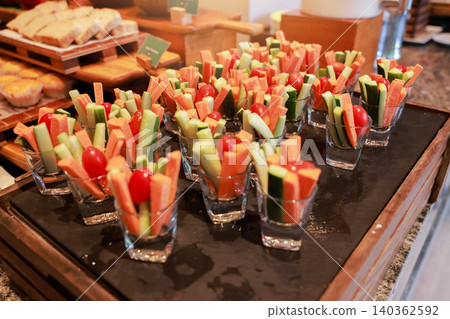 Fresh raw vegetable sticks of carrot and cucumber with cherry tomatoes served in small glass cups on a tray at a hotel breakfast buffet. Healthy vegan diet appetizer snack catering event display. Fresh raw vegetable sticks of carrot and cucumber with cherry tomatoes served in small glass cups on a tray at a hotel breakfast buffet. Healthy vegan diet appetizer snack catering event display. 140362592