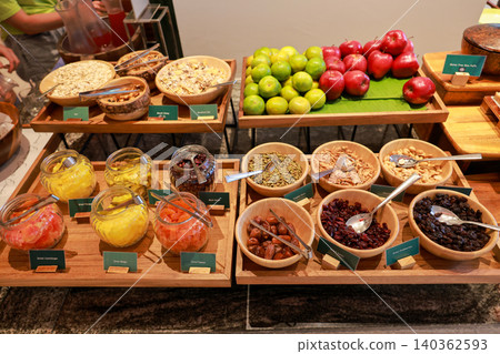Healthy breakfast buffet display at a luxury hotel restaurant featuring fresh apples, oranges, various dried fruits, nuts, seeds, and cereals in wooden bowls and glass jars for morning catering. Healthy breakfast buffet display at a luxury hotel restaurant featuring fresh apples, oranges, various dried fruits, nuts, seeds, and cereals in wooden bowls and glass jars for morning catering. 140362593