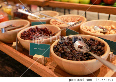 Close up of wooden bowls filled with healthy dried cranberries, dark raisins and mixed nuts at a luxury hotel breakfast buffet station. Sweet organic diet snack for morning catering food display. Close up of wooden bowls filled with healthy dried cranberries, dark raisins and mixed nuts at a luxury hotel breakfast buffet station. Sweet organic diet snack for morning catering food display. 140362594