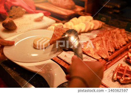 Unrecognizable person using metal tongs to pick up sweet french toast and pancakes onto a white plate at a luxury hotel breakfast buffet. Morning bakery catering service with delicious food. Unrecognizable person using metal tongs to pick up sweet french toast and pancakes onto a white plate at a luxury hotel breakfast buffet. Morning bakery catering service with delicious food. 140362597