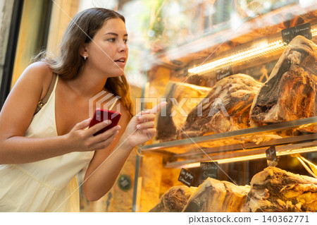 Portrait of young girl with smartphone choosing delicacy of exquisite dry-aged beef at showcase Portrait of young girl with smartphone choosing delicacy of exquisite dry-aged beef at showcase 140362771