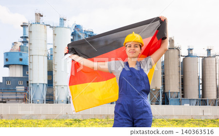 Happy girl in work clothes and hardhat with flag of germany standing in front of industrial scenery Happy girl in work clothes and hardhat with flag of germany standing in front of industrial scenery 140363516