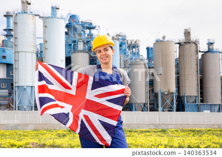Happy girl in work clothes and hardhat with flag of uk standing in front of industrial scenery Happy girl in work clothes and hardhat with flag of uk standing in front of industrial scenery 140363534