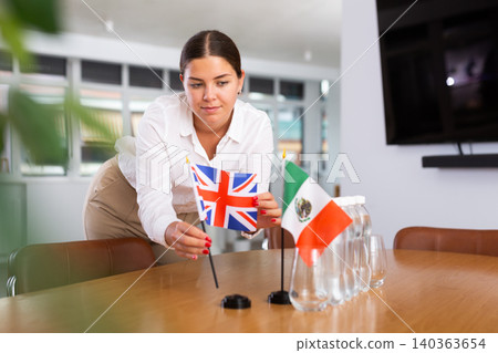 A woman in a white blouse with friendly smile arrangs flags of Mexico and Britain for meeting. A woman in a white blouse with friendly smile arrangs flags of Mexico and Britain for meeting. 140363654