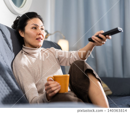 Woman with cup of tea sitting on sofa and watching television Woman with cup of tea sitting on sofa and watching television 140363814