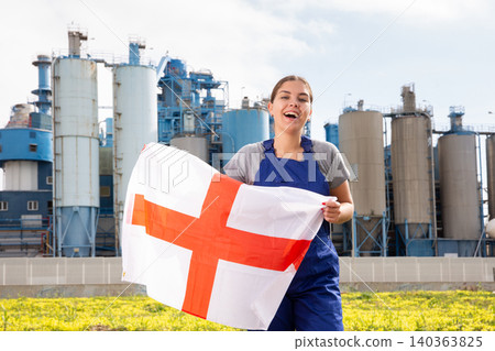 near suburban factory on sunny day, stands gleeful female worker in overalls and holds English flag near suburban factory on sunny day, stands gleeful female worker in overalls and holds English flag 140363825