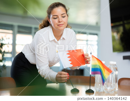 Young woman putting Bahrain and LGBT flags on table in office 140364142