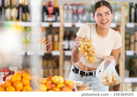 Near counter, young woman client takes out grape from pile of fruits, chooses ripe fruits Near counter, young woman client takes out grape from pile of fruits, chooses ripe fruits 140364505