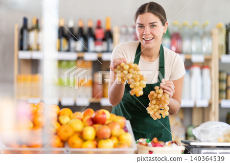 Young woman seller in store is standing near fruit counter, selling grape. Young woman seller in store is standing near fruit counter, selling grape. 140364539