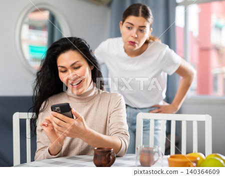 Woman using smartphone at home, her angry friend standing behind and watching 140364609
