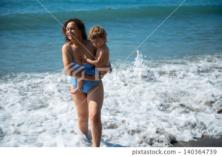 Mother Carrying Toddler in Foamy Beach Waves Mother Carrying Toddler in Foamy Beach Waves 140364739