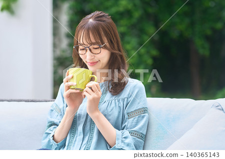 A woman with glasses smiles as she gazes at a mug on a sofa by the window. A woman with glasses smiles as she gazes at a mug on a sofa by the window. 140365143