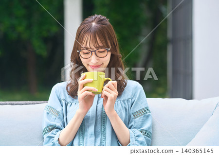 A bespectacled woman enjoying the scent of a yellow mug by the window. 140365145