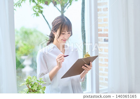 A woman in a white shirt writing on a clipboard by the window. A woman in a white shirt writing on a clipboard by the window. 140365165