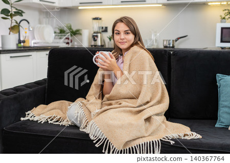 Young woman drinks herbal tea hugs warm mug snuggles in blanket smiles and enjoys peaceful cozy time Young woman drinks herbal tea hugs warm mug snuggles in blanket smiles and enjoys peaceful cozy time 140367764