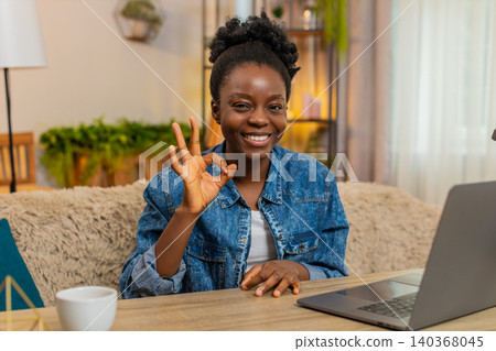 African American young woman smiles showing okay hand gesture while sitting at table on sofa at home 140368045