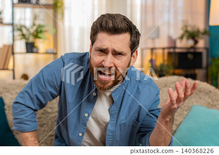 Young man sitting on the sofa at home, squinting at the screen with suspicion, doubting what he sees Young man sitting on the sofa at home, squinting at the screen with suspicion, doubting what he sees 140368226