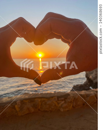 Hands making a heart shape around the setting sun over the ocean at golden hour. Hands making a heart shape around the setting sun over the ocean at golden hour. 140368693