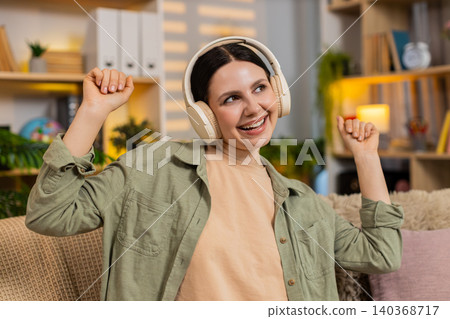 Young woman on home sofa listening to music with wireless headphones smiling in living room indoors Young woman on home sofa listening to music with wireless headphones smiling in living room indoors 140368717