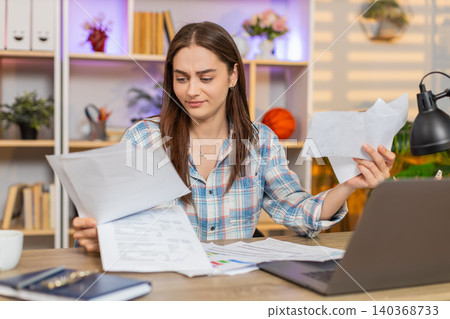 Young woman on home table upset with receipts and documents throwing papers in frustration at laptop Young woman on home table upset with receipts and documents throwing papers in frustration at laptop 140368733