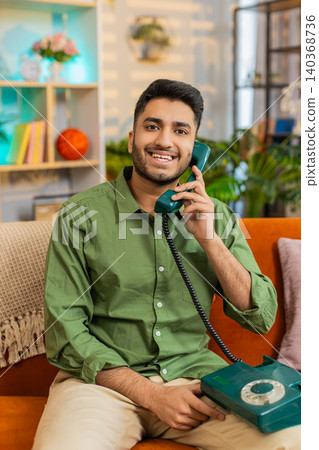 Indian man on home sofa talking on wired phone smiling during happy call with friend about good news Indian man on home sofa talking on wired phone smiling during happy call with friend about good news 140368736
