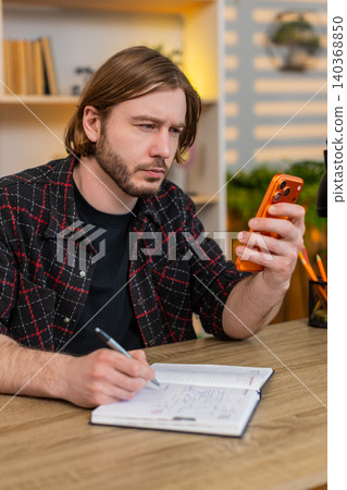 Young man on home table checking smartphone and writing notes for simple personal planning routine Young man on home table checking smartphone and writing notes for simple personal planning routine 140368850