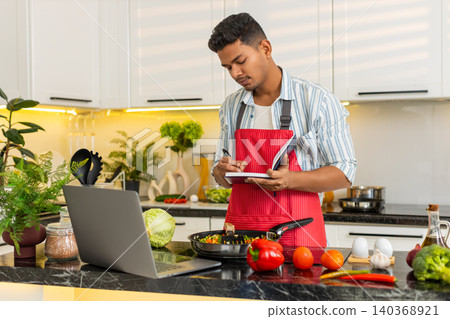 Indian man planning healthy meal notes on laptop and notebook in home kitchen for nutrition lesson Indian man planning healthy meal notes on laptop and notebook in home kitchen for nutrition lesson 140368921