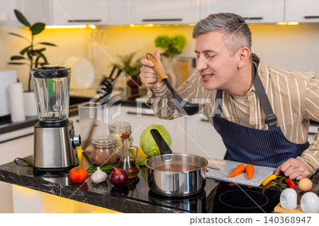 Middle-aged man tasting homemade vegetable soup in kitchen while checking flavor for healthy lunch Middle-aged man tasting homemade vegetable soup in kitchen while checking flavor for healthy lunch 140368947