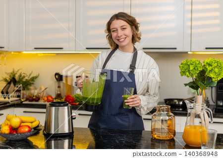 Young woman preparing fresh green smoothie in kitchen at home for healthy breakfast wellness drink 140368948