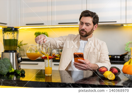 Middle-aged man texting on smartphone while drinking cold orange juice for diet hydration in kitchen Middle-aged man texting on smartphone while drinking cold orange juice for diet hydration in kitchen 140368970