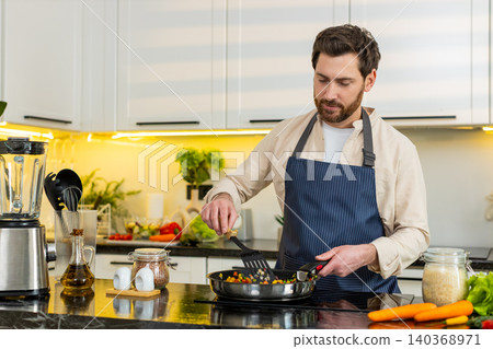Middle-aged man tasting vegetables on skillet pan at home kitchen cooking healthy diet meal on stove 140368971