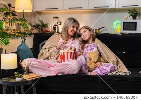 Mother and daughter movie night on sofa with popcorn warm blanket toy bear laugh and enjoy together Mother and daughter movie night on sofa with popcorn warm blanket toy bear laugh and enjoy together 140369007
