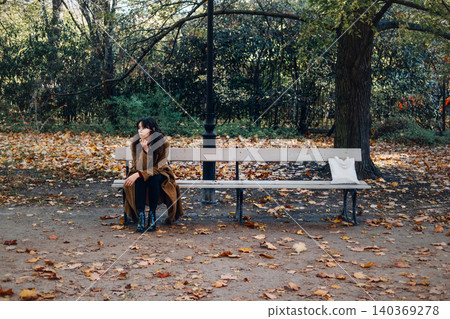 Woman sits thoughtfully on park bench during autumn with white tote bag beside her. Coping with stress, emotional health reflection, managing anxiety outdoors, introverted lifestyle. Woman sits thoughtfully on park bench during autumn with white tote bag beside her. Coping with stress, emotional health reflection, managing anxiety outdoors, introverted lifestyle. 140369278
