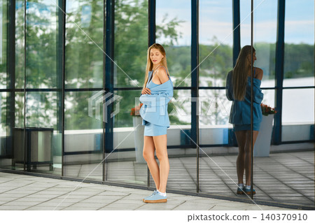 Fashion model poses in total blue outfit outside modern building with large glass windows during daytime Fashion model poses in total blue outfit outside modern building with large glass windows during daytime 140370910