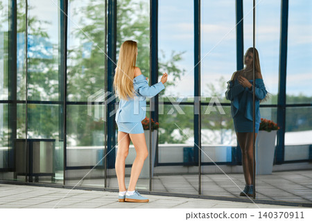 Woman stands near glass wall reflecting her image in urban setting during daytime Woman stands near glass wall reflecting her image in urban setting during daytime 140370911