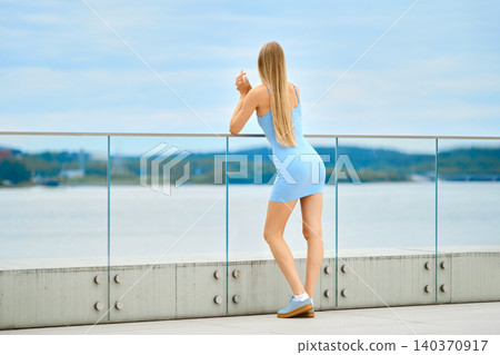Rear view of woman in blue spaghetti strap dress standing at railing overlooking water 140370917