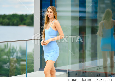 Young woman in blue dress stands by glass railing near water in summer Young woman in blue dress stands by glass railing near water in summer 140370923