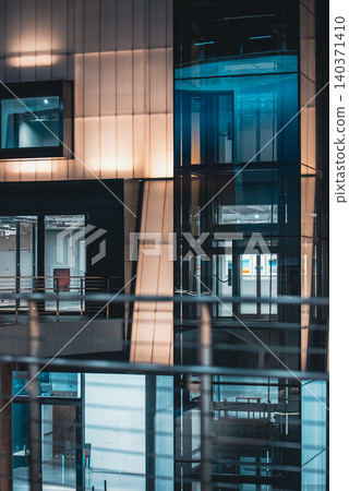 Modern glass elevator shaft with transparent structure and illuminated interior design, Contemporary lift system inside building with reflective surfaces and geometric architecture Modern glass elevator shaft with transparent structure and illuminated interior design, Contemporary lift system inside building with reflective surfaces and geometric architecture 140371410