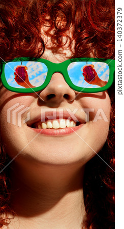 Close up red haired woman in green sunglasses with red drink reflected in lenses on blue sky Close up red haired woman in green sunglasses with red drink reflected in lenses on blue sky 140372379