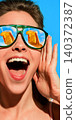 Excited woman in green sunglasses with orange drink reflected in both lenses against blue sky. 140372387