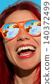 Laughing woman in orange sunglasses with wine poured in mirrored lenses against blue sky. 140372499