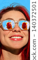 Smiling woman in mirrored sunglasses with red cocktail reflected in both lenses on blue sky background. 140372501