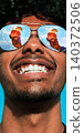 Smiling man in mirrored sunglasses with orange cocktail reflected in lenses against bright blue sky. 140372506