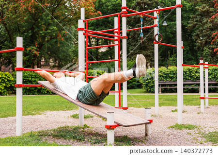 Fit man training his abs and core on an incline bench at an outdoor fitness park. Calisthenics, bodyweight training, functional fitness, and active lifestyle. Fit man training his abs and core on an incline bench at an outdoor fitness park. Calisthenics, bodyweight training, functional fitness, and active lifestyle. 140372773