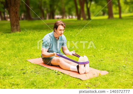 A man performs a stretching exercise with a resistance band on a fitness mat at a park, focusing on his outdoor athlete workout. A man performs a stretching exercise with a resistance band on a fitness mat at a park, focusing on his outdoor athlete workout. 140372777