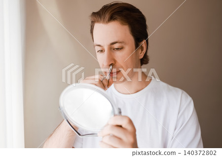Man looking in a mirror during his male grooming routine, using scissors for nose hair removal as part of his daily facial care and self-care concepts. Man looking in a mirror during his male grooming routine, using scissors for nose hair removal as part of his daily facial care and self-care concepts. 140372802