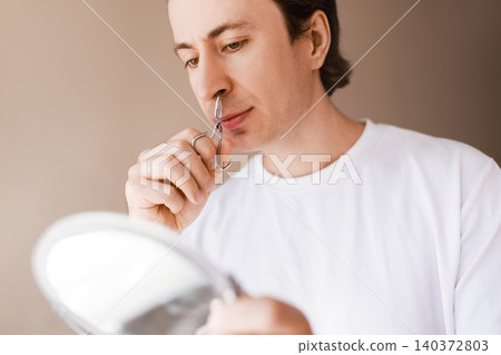 Man trimming nose hair with scissors while looking into a mirror during his male grooming routine, focusing on facial care and self-care concepts. Man trimming nose hair with scissors while looking into a mirror during his male grooming routine, focusing on facial care and self-care concepts. 140372803