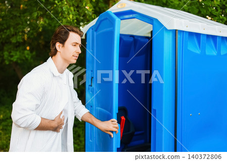Man experiencing an upset stomach and digestive discomfort opening a blue portable outdoor toilet cabin, conveying a gastrointestinal emergency. Man experiencing an upset stomach and digestive discomfort opening a blue portable outdoor toilet cabin, conveying a gastrointestinal emergency. 140372806