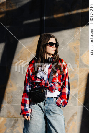 Stylish teen girl wearing sunglasses and headphones, posing against a wall Stylish teen girl wearing sunglasses and headphones, posing against a wall 140374216